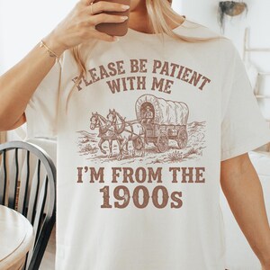 May include: Cream-colored t-shirt with a sepia-toned graphic of a covered wagon pulled by horses. The text on the shirt reads "PLEASE BE PATIENT WITH ME" above the wagon and "I'M FROM THE 1900s" below.