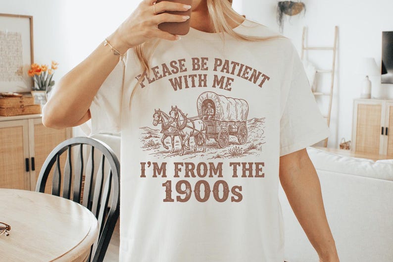 May include: Cream-colored t-shirt with a sepia-toned graphic of a covered wagon pulled by horses. The text on the shirt reads "PLEASE BE PATIENT WITH ME" above the wagon and "I'M FROM THE 1900s" below.