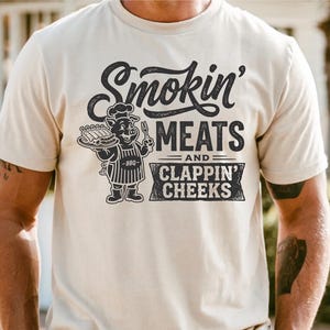 May include: Cream-colored t-shirt with the text "Smokin' MEATS and CLAPPIN' CHEEKS" in a distressed font. A cartoon pig chef holding ribs and a fork is featured in the center. The shirt is short-sleeved.