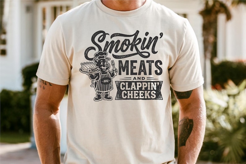 May include: Cream-colored t-shirt with the text "Smokin' MEATS and CLAPPIN' CHEEKS" in a distressed font. A cartoon pig chef holding ribs and a fork is featured in the center. The shirt is short-sleeved.