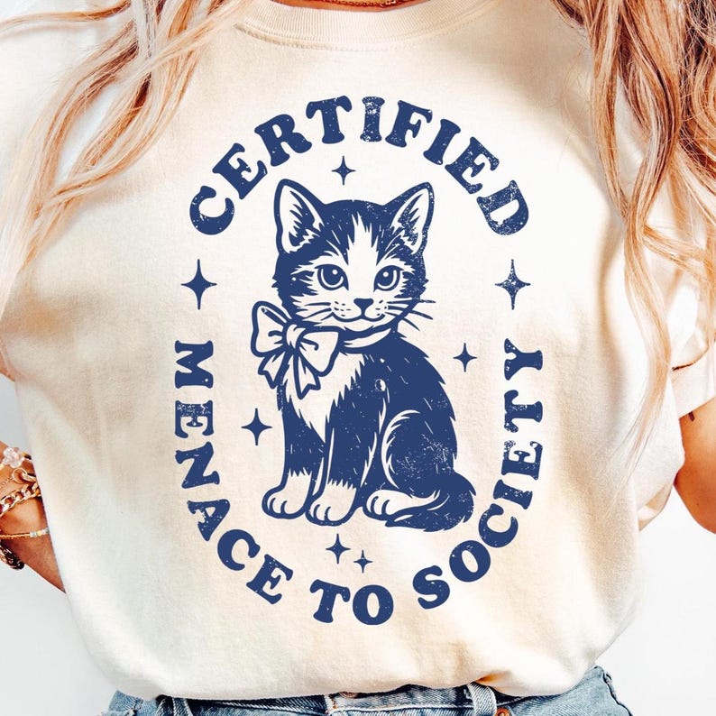 May include: Cream-colored t-shirt with a navy blue graphic of a cat wearing a bow tie, surrounded by the text "CERTIFIED MENACE TO SOCIETY" in a circular design. The design includes star accents.