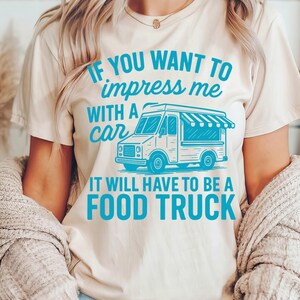 May include: Beige t-shirt with the text "IF YOU WANT TO impress me WITH A car IT WILL HAVE TO BE A FOOD TRUCK" in teal. The graphic features a teal food truck illustration. The shirt is worn by a person.
