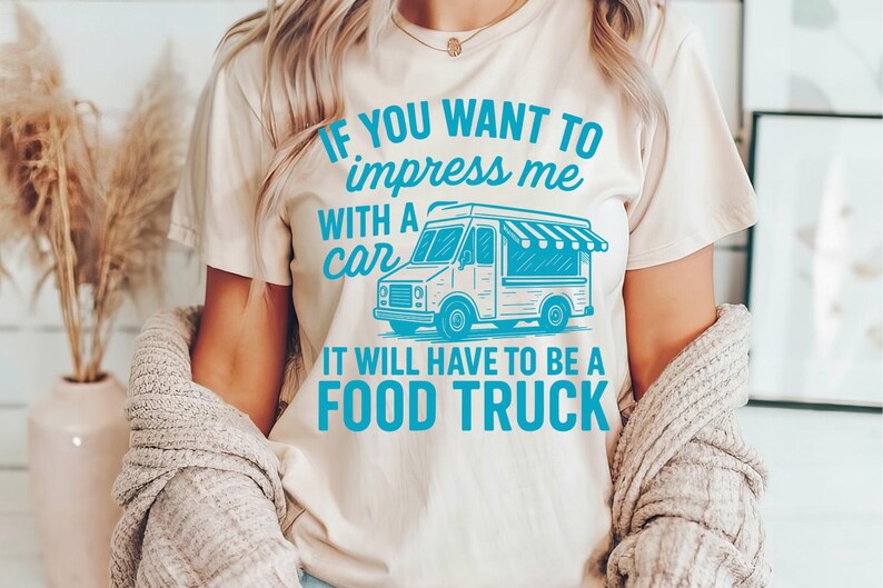 May include: Beige t-shirt with the text "IF YOU WANT TO impress me WITH A car IT WILL HAVE TO BE A FOOD TRUCK" in teal. The graphic features a teal food truck illustration. The shirt is worn by a person.