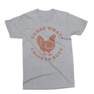 May include: A heather gray t-shirt with a brown chicken graphic and the text "Guess What? Chicken Butt" in a circular design. The shirt is a casual wear item.