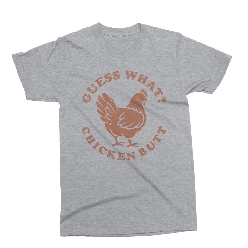 May include: A heather gray t-shirt with a brown chicken graphic and the text "Guess What? Chicken Butt" in a circular design. The shirt is a casual wear item.