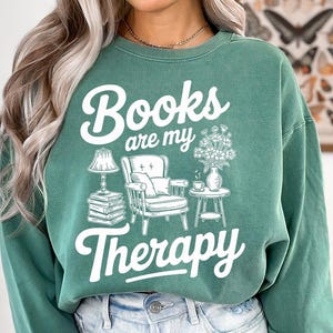 May include: Green sweatshirt with the text "Books are my Therapy" in white script. The graphic includes a lamp, armchair, side table, and flowers. The sweatshirt is a solid green color and has a relaxed fit.