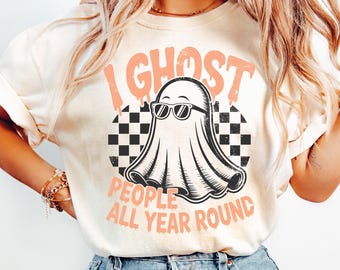 May include: Cream-colored t-shirt featuring a ghost wearing sunglasses, set against a black and white checkered background. The text "I Ghost People All Year Round" is printed in a peach color, creating a playful design.