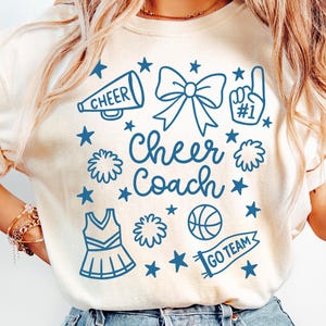 May include: Cream-colored t-shirt with a blue graphic design featuring a megaphone, bow, pom-poms, and the words "Cheer Coach." The design also includes stars, a basketball, and a banner that says "Go Team."