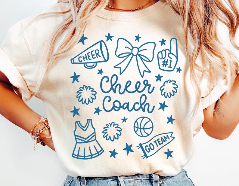 May include: Cream-colored t-shirt with a blue graphic design featuring a megaphone, bow, pom-poms, and the words "Cheer Coach." The design also includes stars, a basketball, and a banner that says "Go Team."
