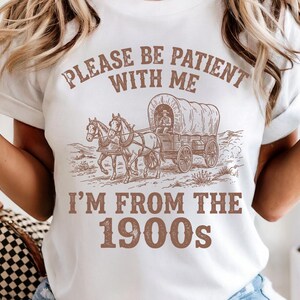 May include: White t-shirt with a sepia-toned graphic of a covered wagon pulled by two horses. The text "PLEASE BE PATIENT WITH ME" arches above the image, with "I'M FROM THE 1900s" below.