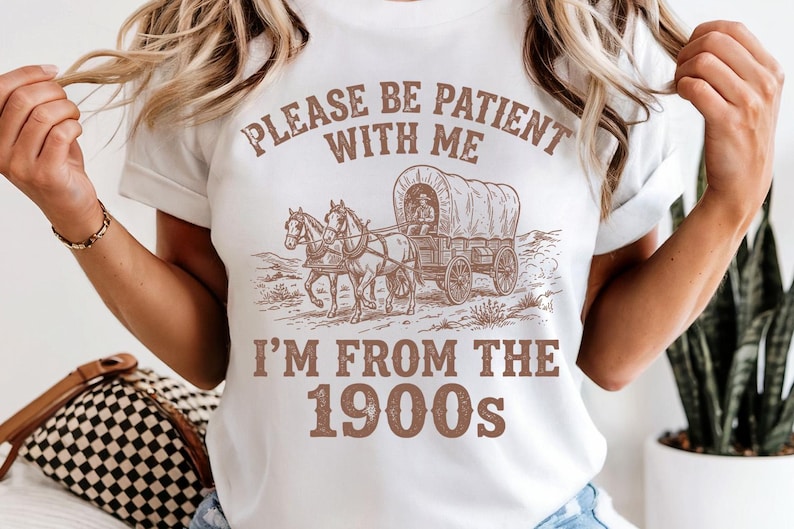 May include: White t-shirt with a sepia-toned graphic of a covered wagon pulled by two horses. The text "PLEASE BE PATIENT WITH ME" arches above the image, with "I'M FROM THE 1900s" below.