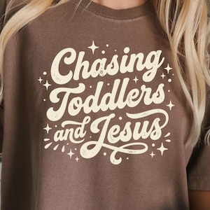May include: A brown t-shirt with the text "Chasing Toddlers and Jesus" in a cream-colored, whimsical font. The text is surrounded by small star graphics. The shirt is made of a soft material.