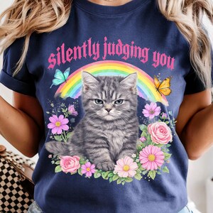 Pu&ograve; includere: T-shirt blu navy con un gatto tabby grigio dall'espressione infastidita, circondato da fiori, farfalle e un arcobaleno. Il testo "silently judging you" &egrave; arcuato sopra il gatto in lettere rosa.
