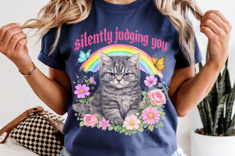 Pu&ograve; includere: T-shirt blu navy con un gatto tabby grigio dall'espressione infastidita, circondato da fiori, farfalle e un arcobaleno. Il testo "silently judging you" &egrave; arcuato sopra il gatto in lettere rosa.