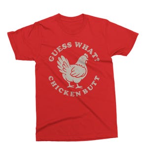 May include: Red t-shirt with a distressed graphic of a chicken and the text "GUESS WHAT? CHICKEN BUTT" in a circular design. The graphic is in a light, off-white color, contrasting with the solid red background of the shirt.
