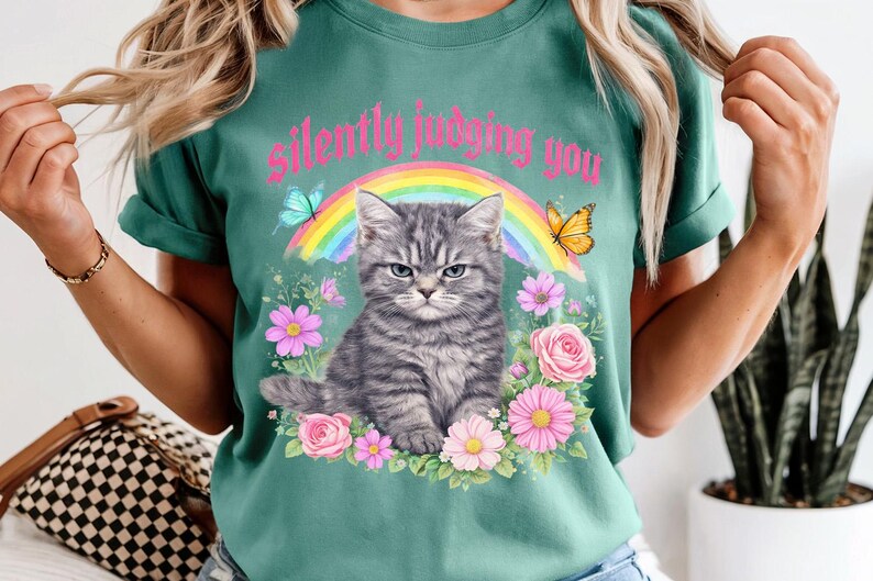 Pu&ograve; includere: T-shirt verde acqua con un'illustrazione di un gatto tabby grigio scontroso, un arcobaleno, farfalle e fiori. Il testo "silently judging you" &egrave; sopra il gatto. La maglietta ha le maniche arrotolate.