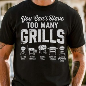May include: Black t-shirt with the text "You Can't Have TOO MANY GRILLS" in white. Below the text are illustrations of different grill types: kettle, offset smoker, barrel, flat top griddle, and classic backyard grill.