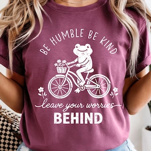 May include: A maroon t-shirt featuring the text "BE HUMBLE BE KIND" and "leave your worries BEHIND" in white. The design includes a white graphic of a frog on a bicycle with flowers. The t-shirt is being worn.