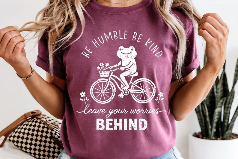 May include: A maroon t-shirt featuring the text "BE HUMBLE BE KIND" and "leave your worries BEHIND" in white. The design includes a white graphic of a frog on a bicycle with flowers. The t-shirt is being worn.