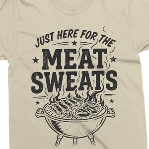 May include: Beige t-shirt with a black graphic. The graphic features the text "JUST HERE FOR THE MEAT SWEATS" above a drawing of a barbecue grill with ribs and steak. The shirt is made of a soft material.