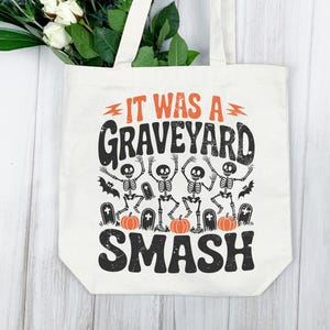 May include: Off-white tote bag with the text "IT WAS A GRAVEYARD SMASH" in black and orange. The design includes dancing skeletons, bats, pumpkins, and tombstones, perfect for Halloween.