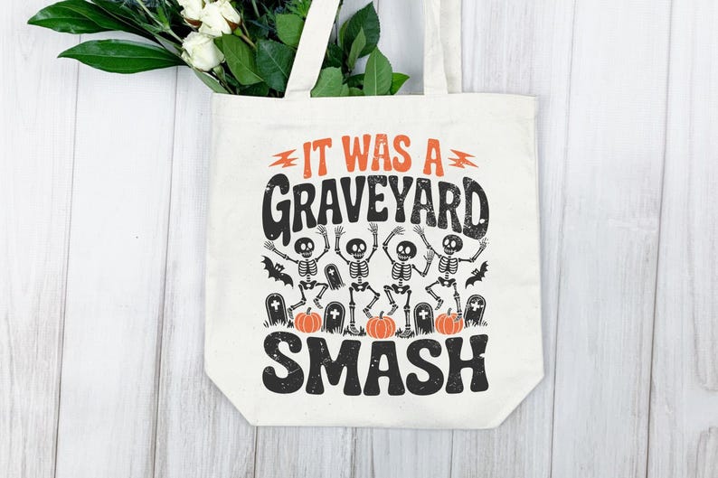 May include: Off-white tote bag with the text "IT WAS A GRAVEYARD SMASH" in black and orange. The design includes dancing skeletons, bats, pumpkins, and tombstones, perfect for Halloween.