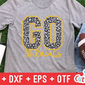 Puede incluir: Una camiseta gris con un dise&ntilde;o de "GO Wildcats" en estampado de leopardo negro y amarillo. La camiseta est&aacute; sobre una superficie de hierba verde con un vaso blanco y una pelota de f&uacute;tbol marr&oacute;n.