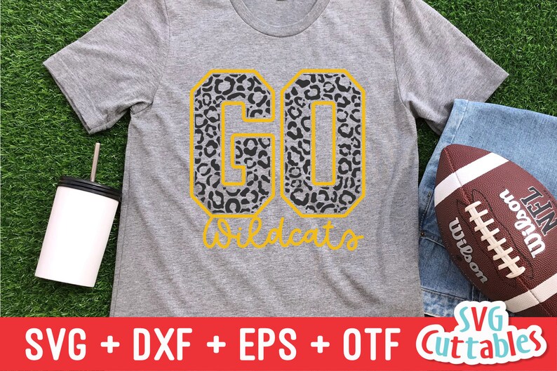 Puede incluir: Una camiseta gris con un dise&ntilde;o de "GO Wildcats" en estampado de leopardo negro y amarillo. La camiseta est&aacute; sobre una superficie de hierba verde con un vaso blanco y una pelota de f&uacute;tbol marr&oacute;n.