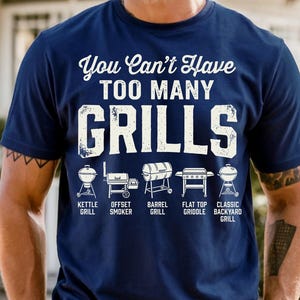 May include: Navy blue t-shirt with the text "You Can't Have TOO MANY GRILLS" in white. Below the text are illustrations of different grill types: kettle, offset smoker, barrel, flat top griddle, and classic backyard grill.