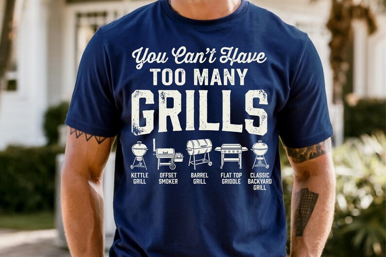 May include: Navy blue t-shirt with the text "You Can't Have TOO MANY GRILLS" in white. Below the text are illustrations of different grill types: kettle, offset smoker, barrel, flat top griddle, and classic backyard grill.
