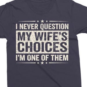 May include: Navy blue t-shirt featuring the white text "I NEVER QUESTION MY WIFE'S CHOICES I'M ONE OF THEM". The text is in a distressed font, arranged in three lines with star accents.