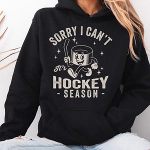 May include: Black hoodie with a front graphic reading "SORRY I CAN'T It's HOCKEY SEASON" in a vintage style. The graphic features a cartoon hockey puck character with a hockey stick. The hoodie has a front pocket and a hood.