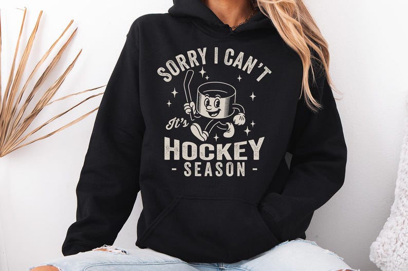 May include: Black hoodie with a front graphic reading "SORRY I CAN'T It's HOCKEY SEASON" in a vintage style. The graphic features a cartoon hockey puck character with a hockey stick. The hoodie has a front pocket and a hood.