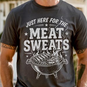 May include: Dark gray t-shirt with the text "JUST HERE FOR THE MEAT SWEATS" in white, along with a graphic of a grill with ribs and steak. The shirt is short-sleeved.