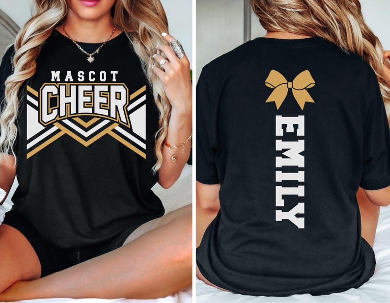 May include: A black t-shirt featuring a front graphic with the text MASCOT CHEER in white and gold block letters above a chevron pattern. The back of the shirt displays a gold cheer bow graphic above the vertical white text EMILY in block letters.