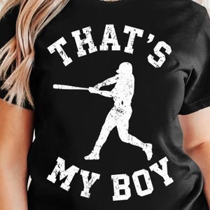 May include: Black t-shirt with the text "THAT'S MY BOY" in white block letters. A white silhouette of a baseball player swinging a bat is centered below the text. The shirt is made of a soft material.