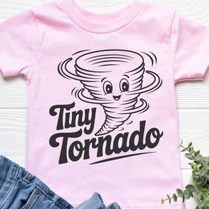 Peut inclure: Un t-shirt rose clair pour enfant avec un dessin noir d'une tornade souriante et les mots "Tiny Tornado". Le t-shirt est en mati&egrave;re douce et est associ&eacute; &agrave; un jean en denim bleu.