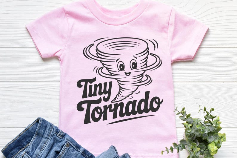 Peut inclure: Un t-shirt rose clair pour enfant avec un dessin noir d'une tornade souriante et les mots "Tiny Tornado". Le t-shirt est en mati&egrave;re douce et est associ&eacute; &agrave; un jean en denim bleu.