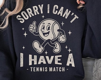 May include: Black sweatshirt with the text "SORRY I CAN'T I HAVE A - TENNIS MATCH -". The design features a cartoon tennis ball character giving a thumbs up and walking, with star accents.