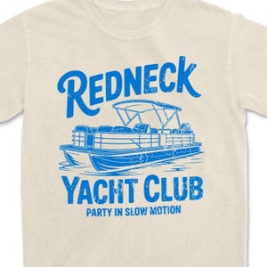 May include: Cream-colored t-shirt with a blue graphic of a pontoon boat and the words "Redneck Yacht Club" above and "Party in Slow Motion" below. The design is centered on the front of the shirt.