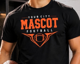 May include: A black short-sleeved t-shirt featuring the orange text "YOUR CITY MASCOT FOOTBALL" above a stylized orange graphic of a football centered within a geometric frame. The design is printed on the chest of the crew neck garment.