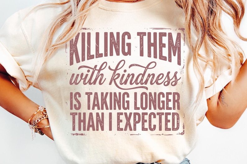 K&ouml;nnte beinhalten: Cremefarbenes T-Shirt mit dem Spruch "KILLING THEM with kindness IS TAKING LONGER THAN I EXPECTED" in einer verblassten, kastanienbraunen Schrift. Das Shirt hat einen Rundhalsausschnitt und kurze &Auml;rmel.