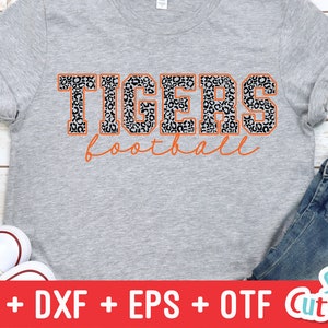Puede incluir: Una camiseta gris con un dise&ntilde;o de estampado de leopardo en blanco y negro. El texto "TIGERS football" est&aacute; impreso en naranja en la camiseta.