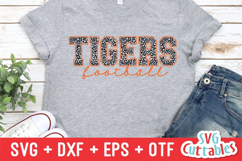 Puede incluir: Una camiseta gris con un dise&ntilde;o de estampado de leopardo en blanco y negro. El texto "TIGERS football" est&aacute; impreso en naranja en la camiseta.