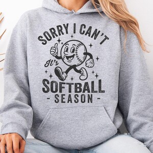 Op de afbeelding: Een lichtgrijze hoodie met een voorzak en een capuchon met trekkoord. De voorkant heeft een zwarte afbeelding van een honkbal met een gezicht, armen en benen, met de tekst "SORRY I CAN'T It's SOFTBALL SEASON".