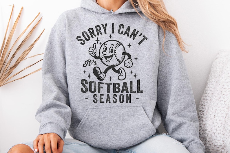 Op de afbeelding: Een lichtgrijze hoodie met een voorzak en een capuchon met trekkoord. De voorkant heeft een zwarte afbeelding van een honkbal met een gezicht, armen en benen, met de tekst "SORRY I CAN'T It's SOFTBALL SEASON".