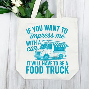 May include: Off-white tote bag with the text "IF YOU WANT TO impress me WITH A car IT WILL HAVE TO BE A FOOD TRUCK" in teal. The bag features a teal illustration of a food truck. White flowers and greenery are visible.
