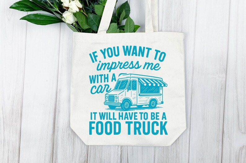 May include: Off-white tote bag with the text "IF YOU WANT TO impress me WITH A car IT WILL HAVE TO BE A FOOD TRUCK" in teal. The bag features a teal illustration of a food truck. White flowers and greenery are visible.