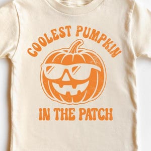May include: Cream-colored t-shirt featuring an orange graphic of a pumpkin wearing sunglasses. The text above the pumpkin reads "COOLEST PUMPKIN" and below it reads "IN THE PATCH".