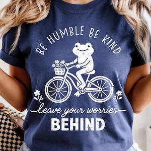 May include: Navy blue t-shirt featuring a white frog on a bicycle, with a basket of flowers. The text says "Be Humble Be Kind" and "Leave Your Worries Behind". A relaxed fit, casual tee.